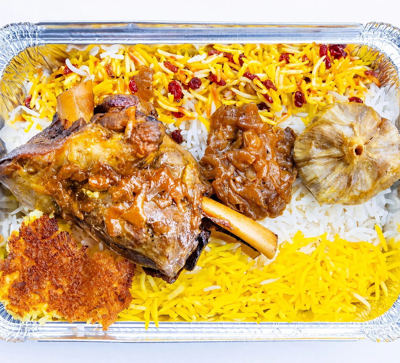 Braised lamb shank in tomato sauce with a roasted garlic bulb, saffron rice with barberries and crispy tahdig in a foil tray at AFRA Grill, Saskatoon
