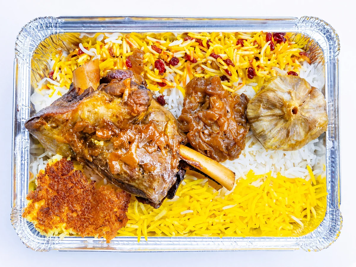 Braised lamb shank in tomato sauce with a roasted garlic bulb, saffron rice with barberries and crispy tahdig in a foil tray at AFRA Grill, Saskatoon